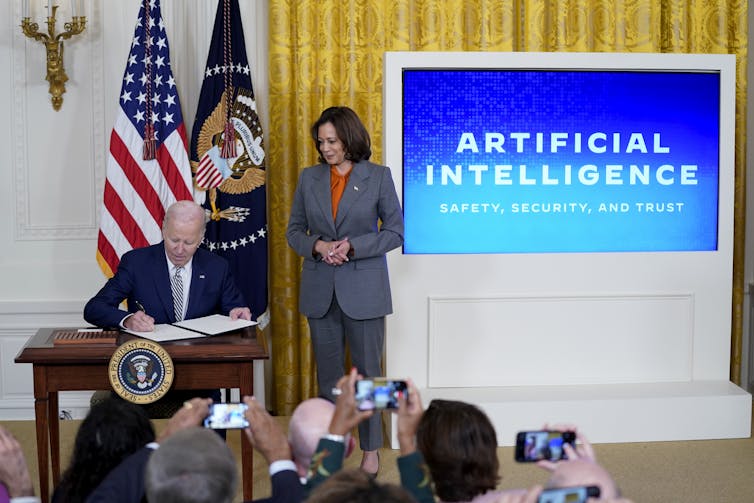 a man sits at a desk writing on a piece of paper as a woman looks on